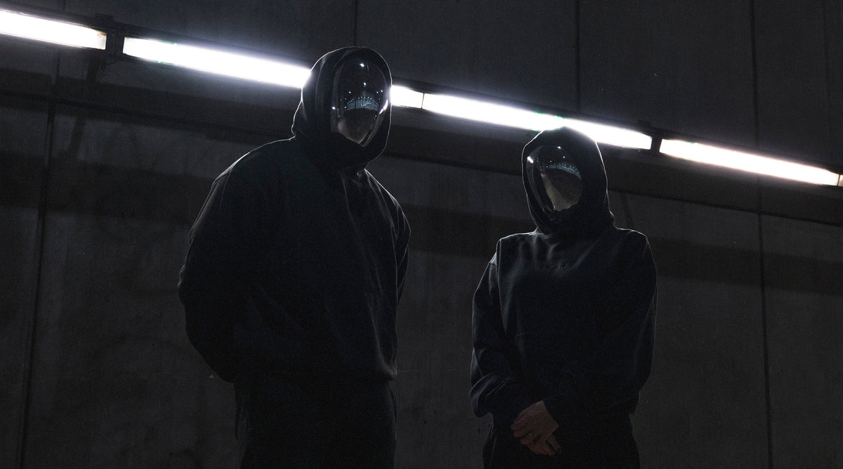 two people in FOFF masks and black hoodies in a dark industrial space under strip lights, collection 02 campaign